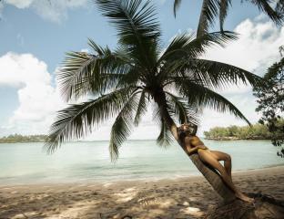 A woman in a bikini lounging against a palm tree on the beach