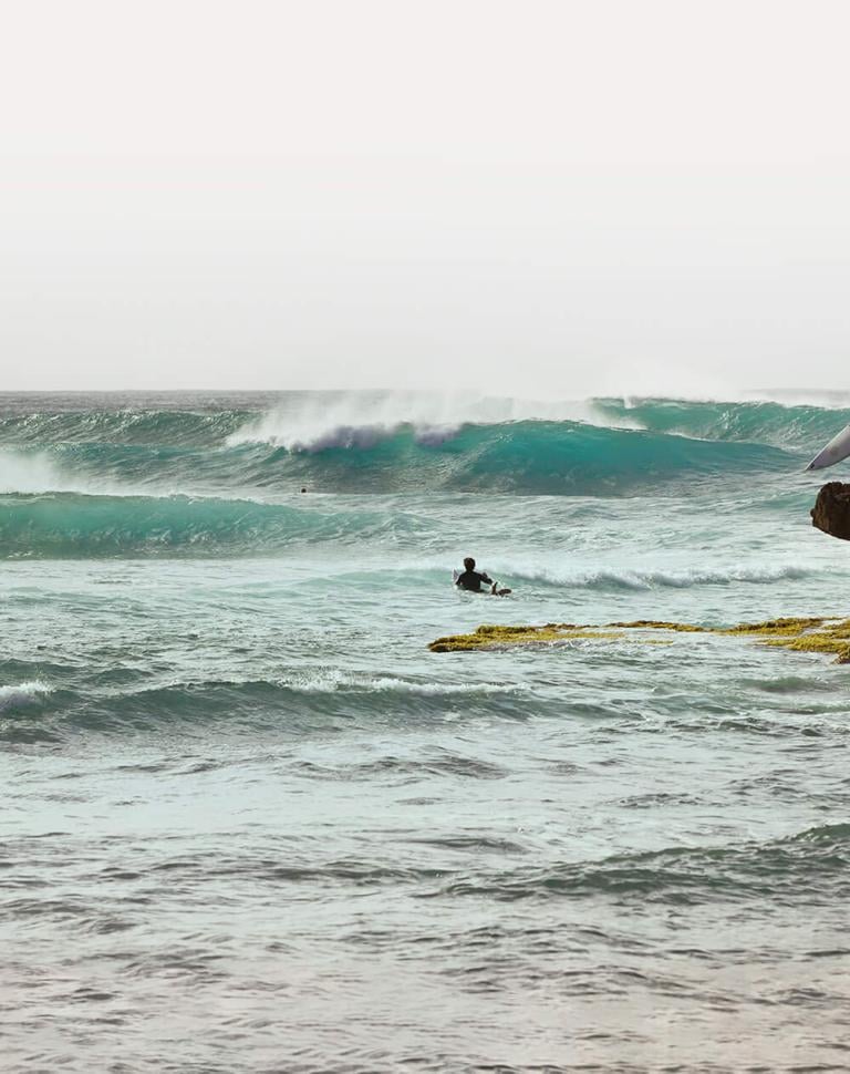 A man standing on a rocky shoreline holds a surfboard while watching people surf in the distance