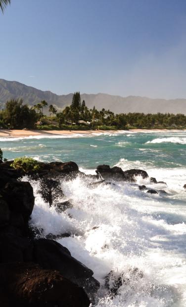 The coast of tropical Hawaii with mountains in the background and crashing waves on a beach.